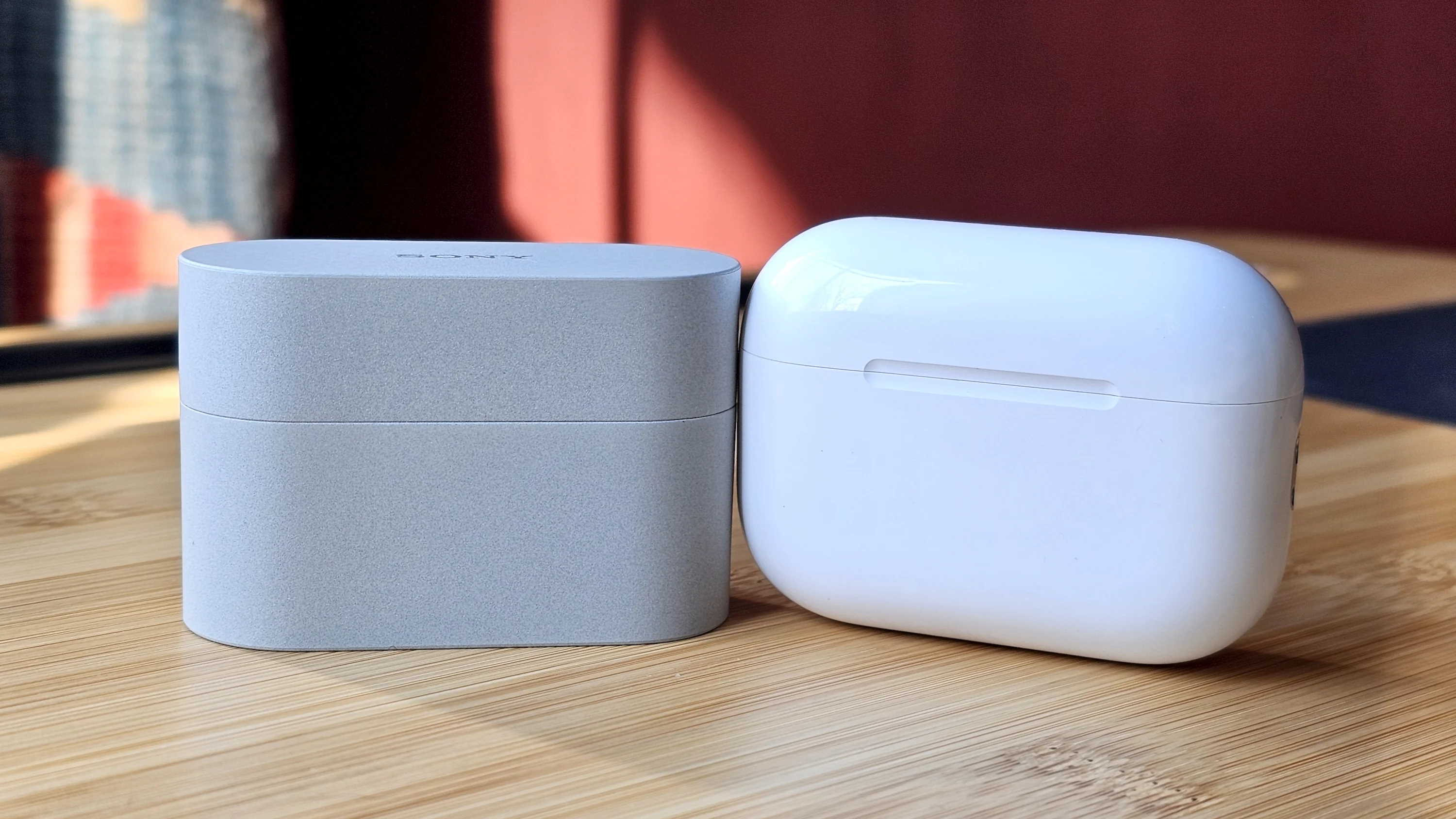 Sony WF-1000XM6 earbuds next to the Apple AirPods Pro 3, on a wooden table, in a versus-style format 
