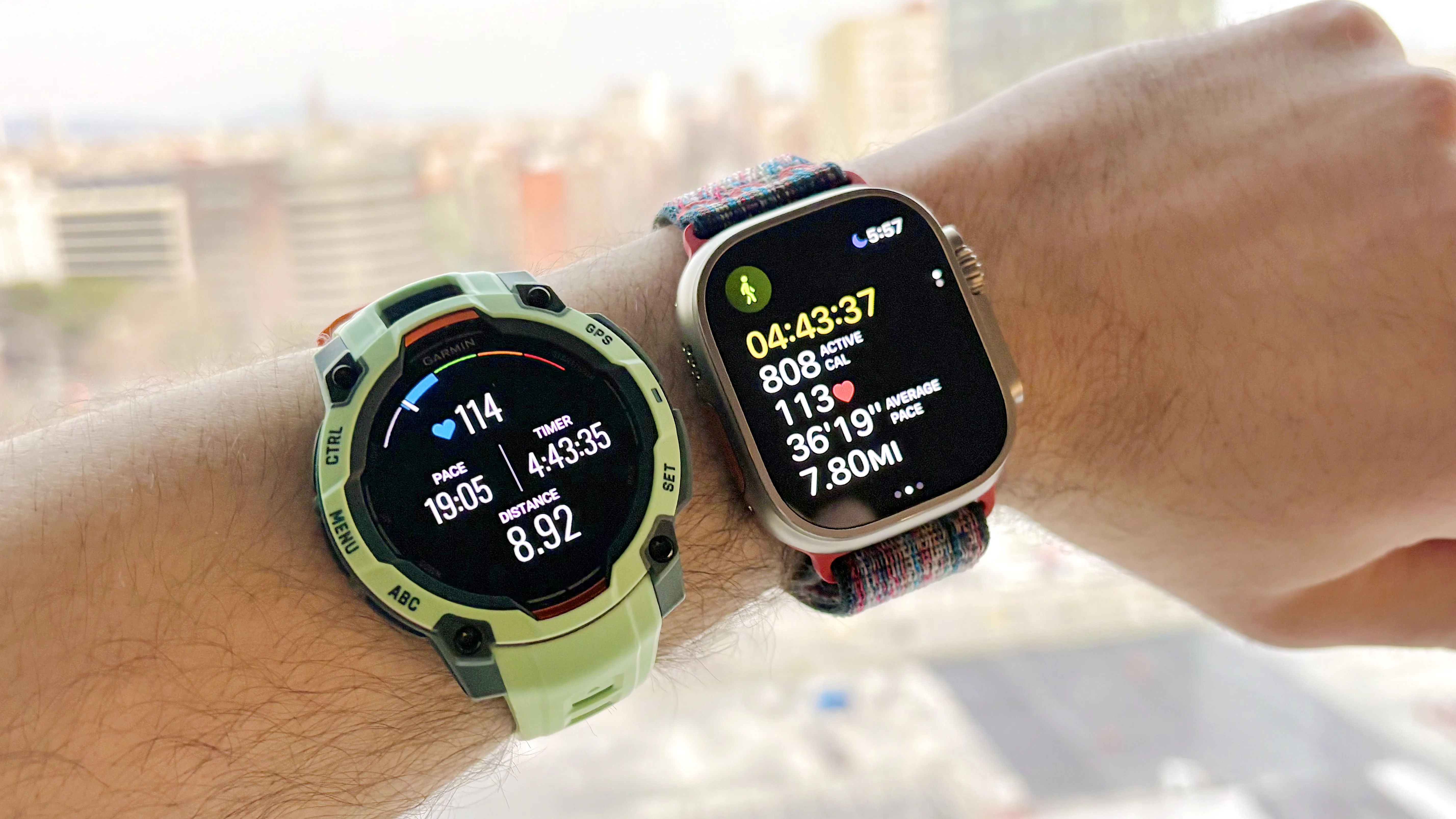 Close-up of the Garmin Instinct 3 next to the Apple Watch Ultra 3 with the Barcelona skyline in the distance
